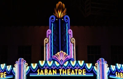 The Saban Theatre's neon marquee glowing blue, pink and gold at night, with the theatre's name spelled out across the front