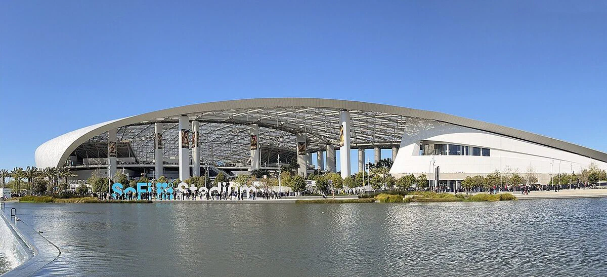 SoFi Stadium's curved white roof and glass sides across the lake at Hollywood Park in Inglewood on a clear day